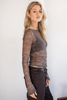 Woman wearing a TIC TOC TC31606 MIRELLE grey stretch sheer lace long sleeve top featuring a wide boat neckline, fitted sleeves, and see-through lace fabric, and brown cargo pants against a white background.