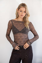 Woman wearing a TIC TOC TC31606 MIRELLE grey stretch sheer lace long sleeve top featuring a wide boat neckline, fitted sleeves, and see-through lace fabric, and brown cargo pants against a white background.