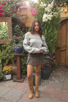 Woman wearing the TIC TOC TC31658 HAZE grey knit off the shoulder sweater featuring a foldover neckline, ribbed cuffs, and relaxed fit paired with a brown denim skirt and tan boots outdoors.