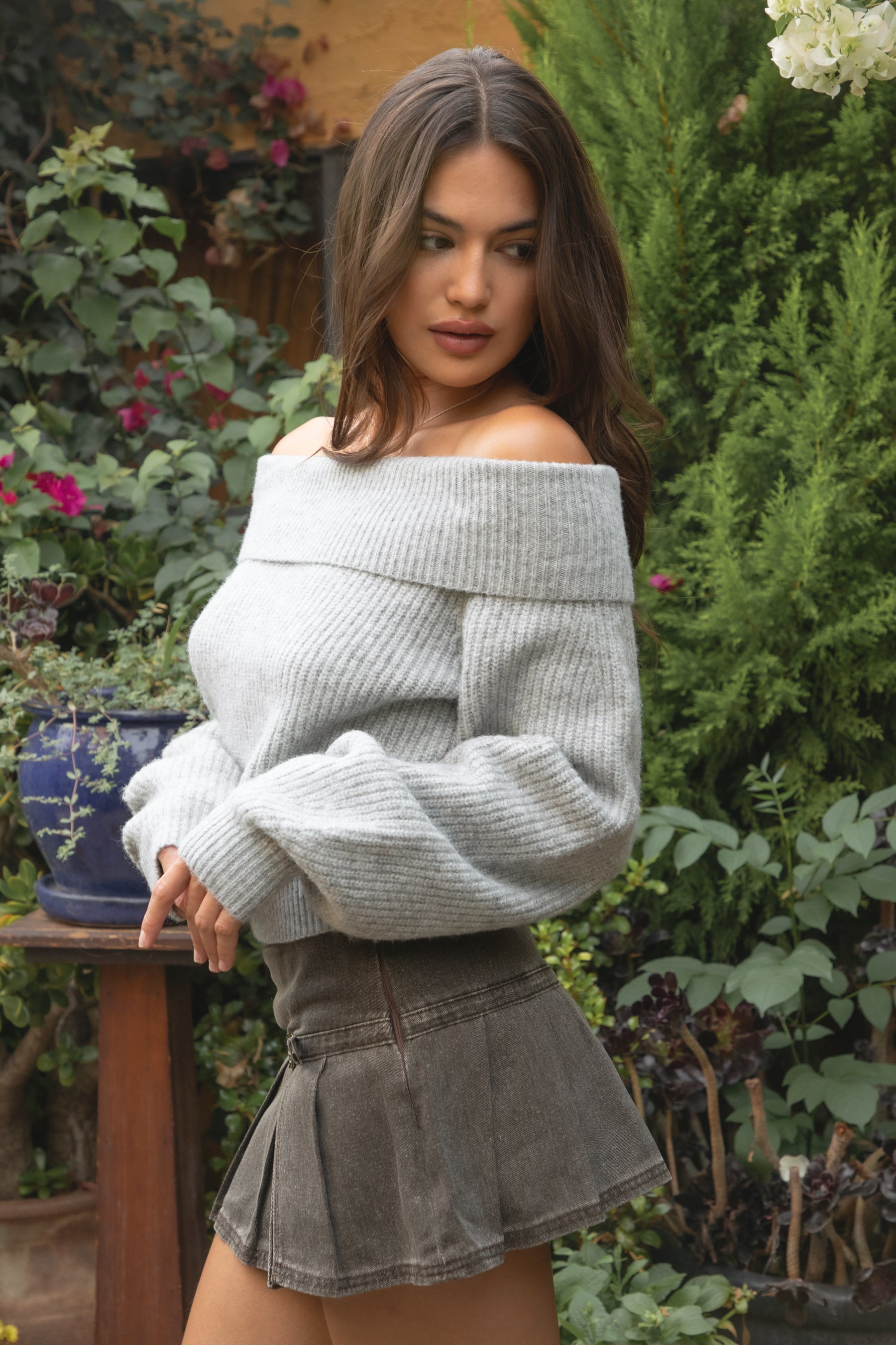 Woman wearing the TIC TOC TC31658 HAZE grey knit off the shoulder sweater featuring a foldover neckline, ribbed cuffs, and relaxed fit paired with a brown denim skirt outdoors.