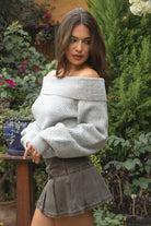 Woman wearing the TIC TOC TC31658 HAZE grey knit off the shoulder sweater featuring a foldover neckline, ribbed cuffs, and relaxed fit paired with a brown denim skirt outdoors.