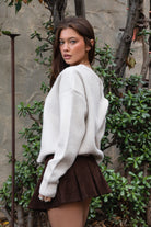 Woman wearing TIC TOC TC31667 BRIDLEWOOD intarsia horse knit sweater with off-the-shoulder ribbed neckline and relaxed fit and brown skirt standing outdoors.