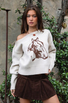 Woman wearing TIC TOC TC31667 BRIDLEWOOD intarsia horse knit sweater with off-the-shoulder ribbed neckline and relaxed fit and brown skirt standing outdoors.