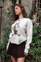 Woman wearing TIC TOC TC31667 BRIDLEWOOD intarsia horse knit sweater with off-the-shoulder ribbed neckline and relaxed fit and brown skirt standing outdoors.