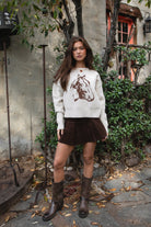 Woman wearing TIC TOC TC31667 BRIDLEWOOD intarsia horse knit sweater with off-the-shoulder ribbed neckline and relaxed fit and brown skirt standing outdoors.