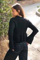 Woman wearing TIC TOC TC31678 LETTIE black sweater knit V-neck cardigan with flared sleeves, cropped peplum hem, and ribbed stretch fabric with dark denim jeans in an outdoor setting. 