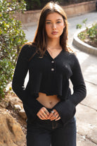 Woman wearing TIC TOC TC31678 LETTIE black sweater knit V-neck cardigan with flared sleeves, cropped peplum hem, and ribbed stretch fabric with dark denim jeans in an outdoor setting. 