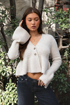 Woman wearing TIC TOC TC31678 LETTIE oatmeal sweater knit V-neck cardigan with flared sleeves, cropped peplum hem, and ribbed stretch fabric with dark denim jeans in an outdoor setting
