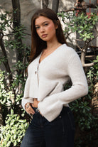 Woman wearing TIC TOC TC31678 LETTIE oatmeal sweater knit V-neck cardigan with flared sleeves, cropped peplum hem, and ribbed stretch fabric with dark denim jeans in an outdoor setting