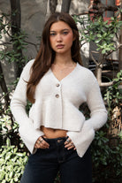 Woman wearing TIC TOC TC31678 LETTIE oatmeal sweater knit V-neck cardigan with flared sleeves, cropped peplum hem, and ribbed stretch fabric with dark denim jeans in an outdoor setting