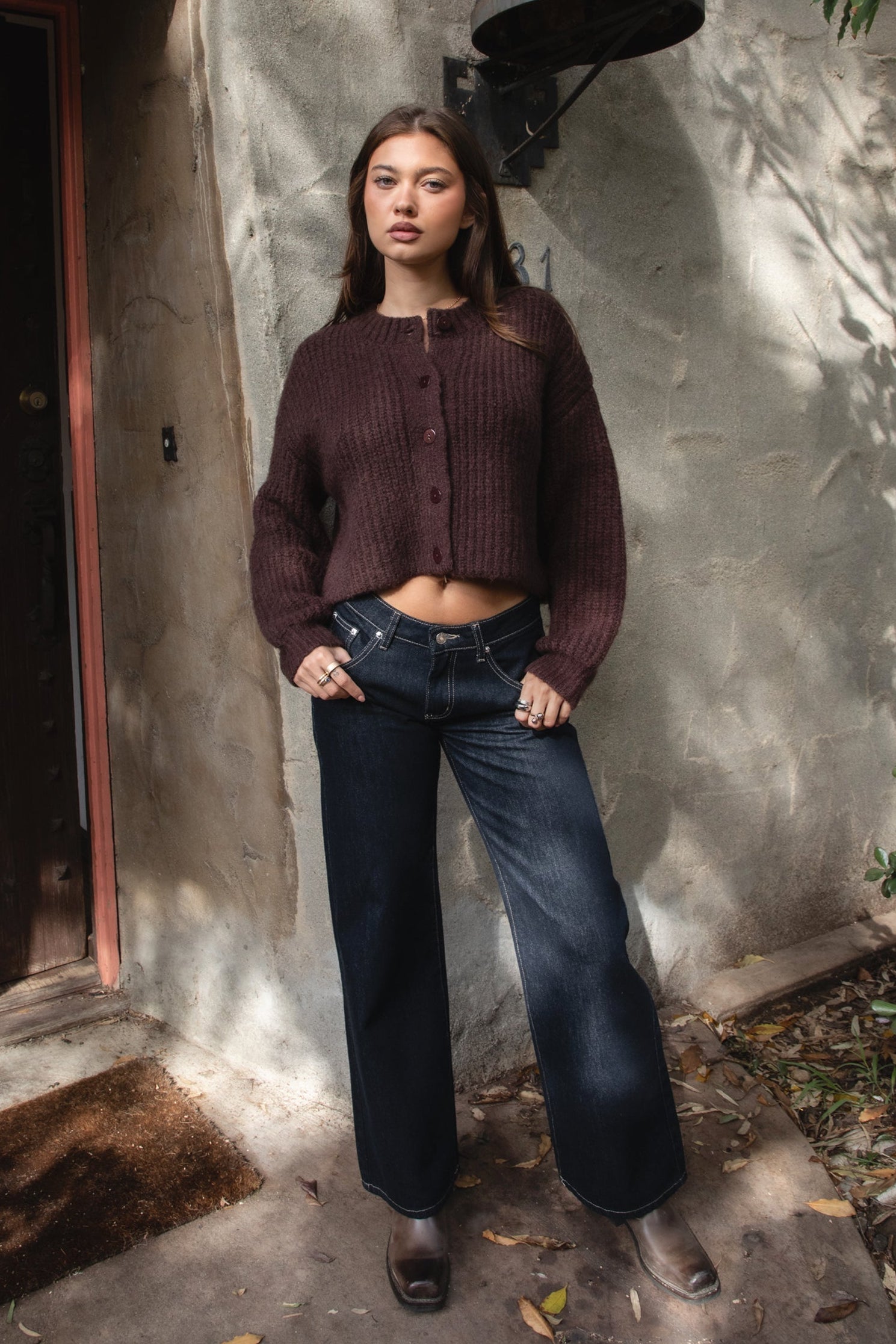 Woman wearing TIC TOC TF31833 ISOLDE brown wool-blend button down cardigan with boxy cropped fit, ribbed trim, and marled knit texture paired with dark jeans and boots against a textured wall.