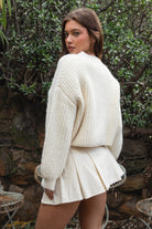 Woman wearing TIC TOC TF31833 ISOLDE cream wool-blend button down cardigan with boxy cropped fit, ribbed trim, and marled knit texture paired with a skirt outdoors with greenery in the background