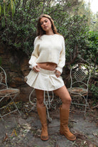 Woman wearing TIC TOC TF31833 ISOLDE cream wool-blend button down cardigan with boxy cropped fit, ribbed trim, and marled knit texture paired with a skirt adn tan boots outdoors with greenery in the background