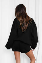 Person wearing a black oversized sweater and shorts set against a white curtain background