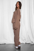 Woman wearing a brown tracksuit standing against a white curtain.
