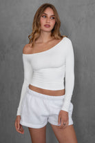 Woman wearing TIC TOC TH30538 AUDREE lightweight one-shoulder long sleeve top in slim fit stretch fabric in color white paired with fleece shorts. 