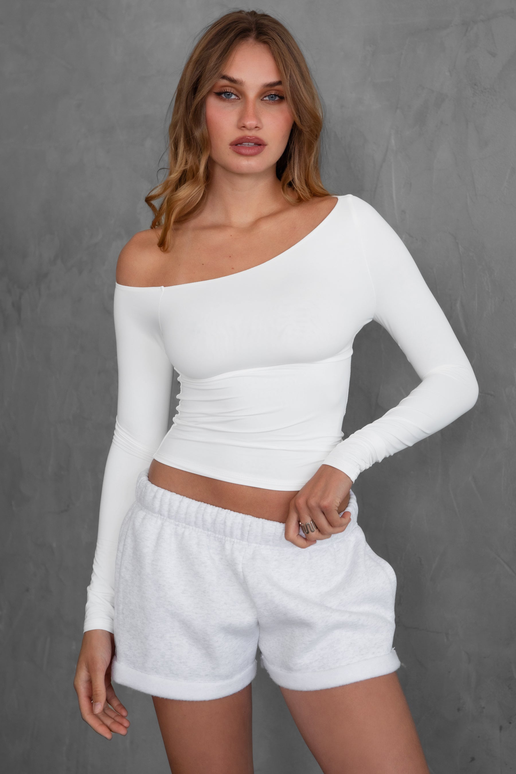 Woman wearing TIC TOC TH30538 AUDREE lightweight one-shoulder long sleeve top in slim fit stretch fabric in color white paired with fleece shorts. 
