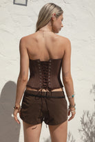 Model wearing the TIC TOC TH30975 LAUREL brown mesh strapless bustier corset top featuring ruffle trim edges, underwire cups, and lace-up back paired with brown suede shorts, standing against a wall