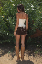 Model wearing the TIC TOC TH30975 LAUREL cream mesh strapless bustier corset top featuring ruffle trim edges, underwire cups, and lace-up back paired with brown suede shorts, standing against  greenery