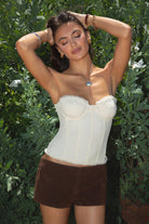 Model wearing the TIC TOC TH30975 LAUREL cream mesh strapless bustier corset top featuring ruffle trim edges, underwire cups, and lace-up back paired with brown suede shorts, standing against  greenery