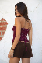 Model wearing the TIC TOC TH30975 LAUREL wine mesh strapless bustier corset top featuring ruffle trim edges, underwire cups, and lace-up back paired with brown suede shorts, standing against a wall