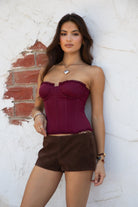 Model wearing the TIC TOC TH30975 LAUREL wine mesh strapless bustier corset top featuring ruffle trim edges, underwire cups, and lace-up back paired with brown suede shorts, standing against a wall