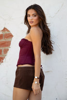 Model wearing the TIC TOC TH30975 LAUREL wine mesh strapless bustier corset top featuring ruffle trim edges, underwire cups, and lace-up back paired with brown suede shorts, standing against a wall
