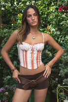 Woman wearing the TIC TOC TH31487 CHARLOTTE floral poplin print corset top featuring lace trim, adjustable straps, bow detail, and lace-up back paired with brown suede shorts in an outdoor green setting. 