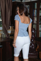 Woman wearing TIC TOC TH31535 SAILYN pinstripe poplin short sleeve top with scoop neckline, puff sleeves, and button down detail paired with white capri pants in a library. 
