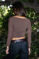 Woman wearing TIC TOC TH31575 MARCELLE brown asymmetric off the shoulder long sleeve top with wide boat neckline and ruched waist paired with dark denim standing in front of green foliage.