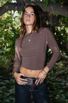 Woman wearing TIC TOC TH31575 MARCELLE brown asymmetric off the shoulder long sleeve top with wide boat neckline and ruched waist paired with dark denim standing in front of green foliage.