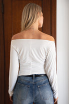Woman wearing a white off-shoulder top and blue jeans standing in front of a wooden door.