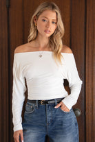 Woman wearing a white off-shoulder top and blue jeans against a wooden door background