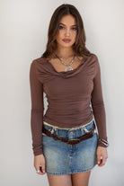 Woman wearing the TIC TOC TH31664 VELARI brown cowl neck long sleeve top featuring a draped neckline, off-the-shoulder fit, and side ruching detail paired with a denim mini skirt