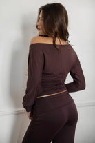 Woman wearing the TIC TOC TH31856BA SORA brown asymmetric off-the-shoulder long sleeve top featuring a slouchy cropped fit, wide boat neckline, and soft stretch fabric with matching pants against a white walls 