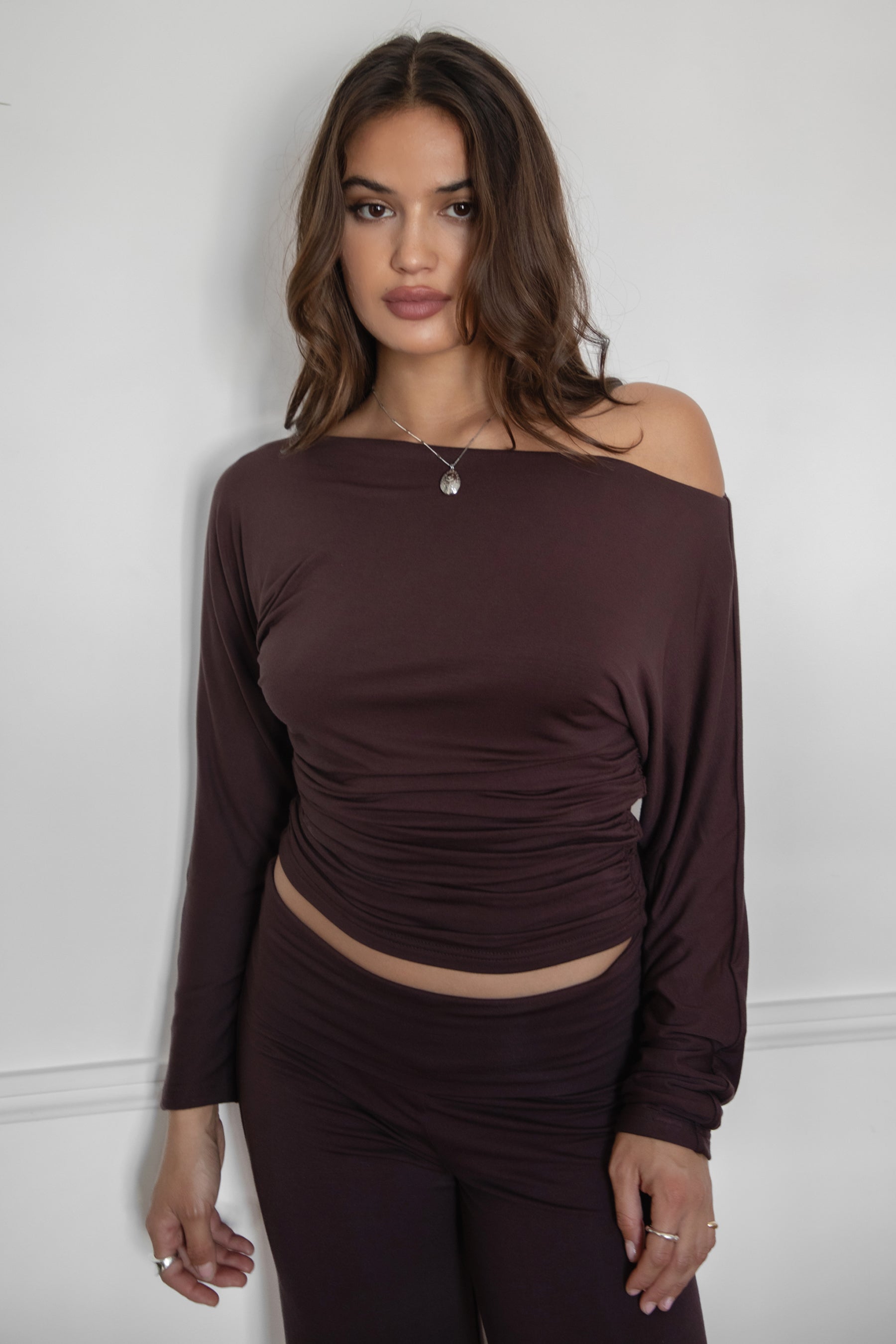 Woman wearing the TIC TOC TH31856BA SORA brown asymmetric off-the-shoulder long sleeve top featuring a slouchy cropped fit, wide boat neckline, and soft stretch fabric with matching pants against a white walls 
