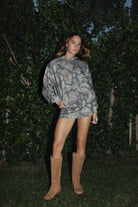 Woman wearing TIC TOC TJ31428 ELK woodland camo oversized fleece hoodie with kangaroo pocket and ribbed cuffs in neutral earth tones with matching bottoms and tan boots in front of a dark background