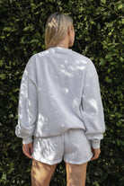 Woman wearing the TIC TOC TJ31656 LANI ash cloud fleece crew neck pullover sweatshirt featuring dropped shoulders, ribbed trim, and cozy fleece fabric with the matching bottoms  against a green leafy background.