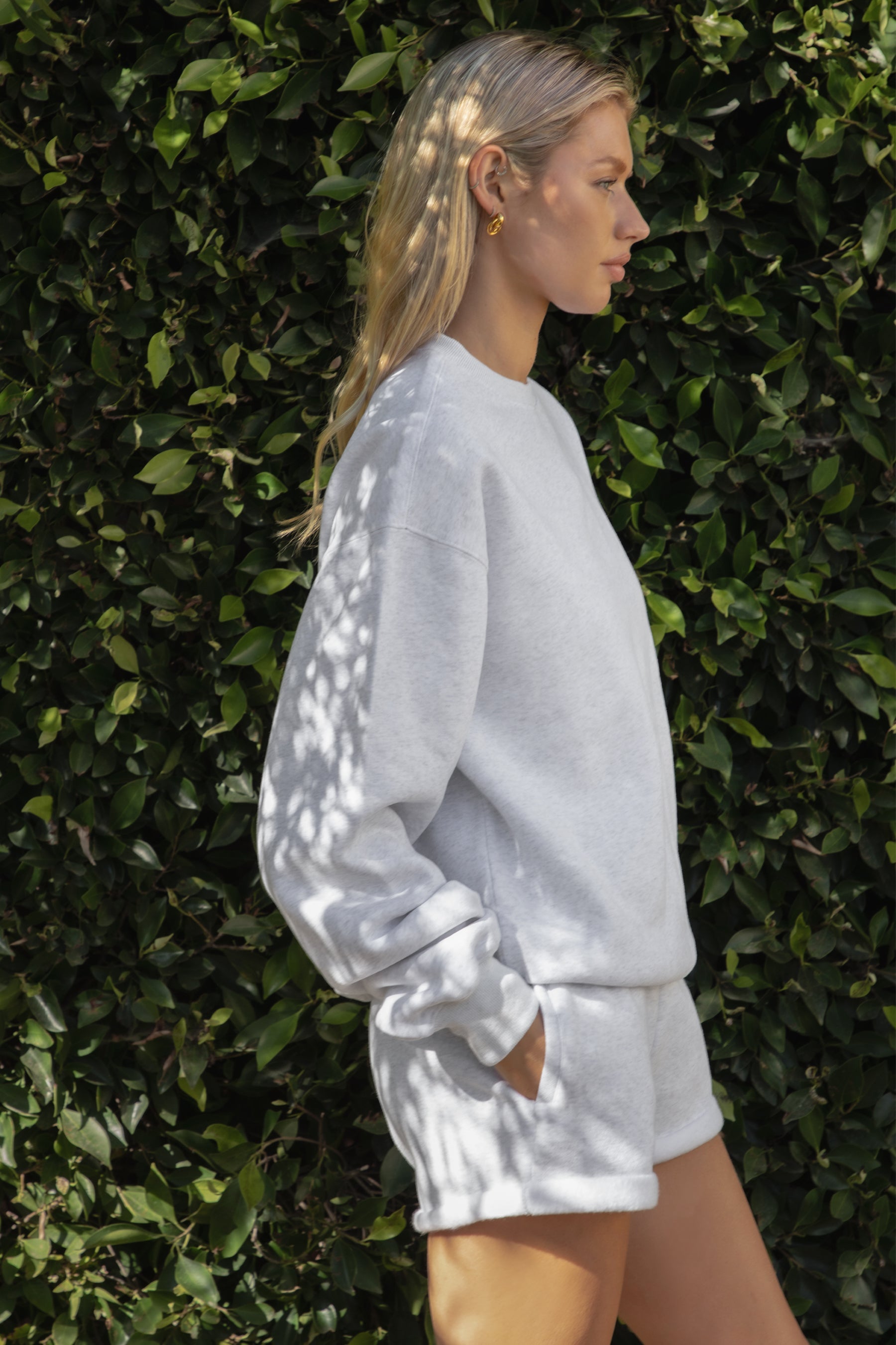 Woman wearing the TIC TOC TJ31656 LANI ash cloud fleece crew neck pullover sweatshirt featuring dropped shoulders, ribbed trim, and cozy fleece fabric with the matching bottoms  against a green leafy background.