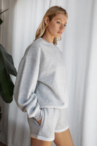 Woman wearing the TIC TOC TJ31656 LANI heather grey cloud fleece crew neck pullover sweatshirt featuring dropped shoulders, ribbed trim, and cozy fleece fabric with the matching bottoms .
