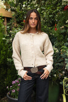 Woman wearing TIC TOC TV31637 MARLO oatmeal sweater knit button down cardigan with ribbed cuffs and dropped shoulders in cozy marled fabric paired with dark denim in a garden setting