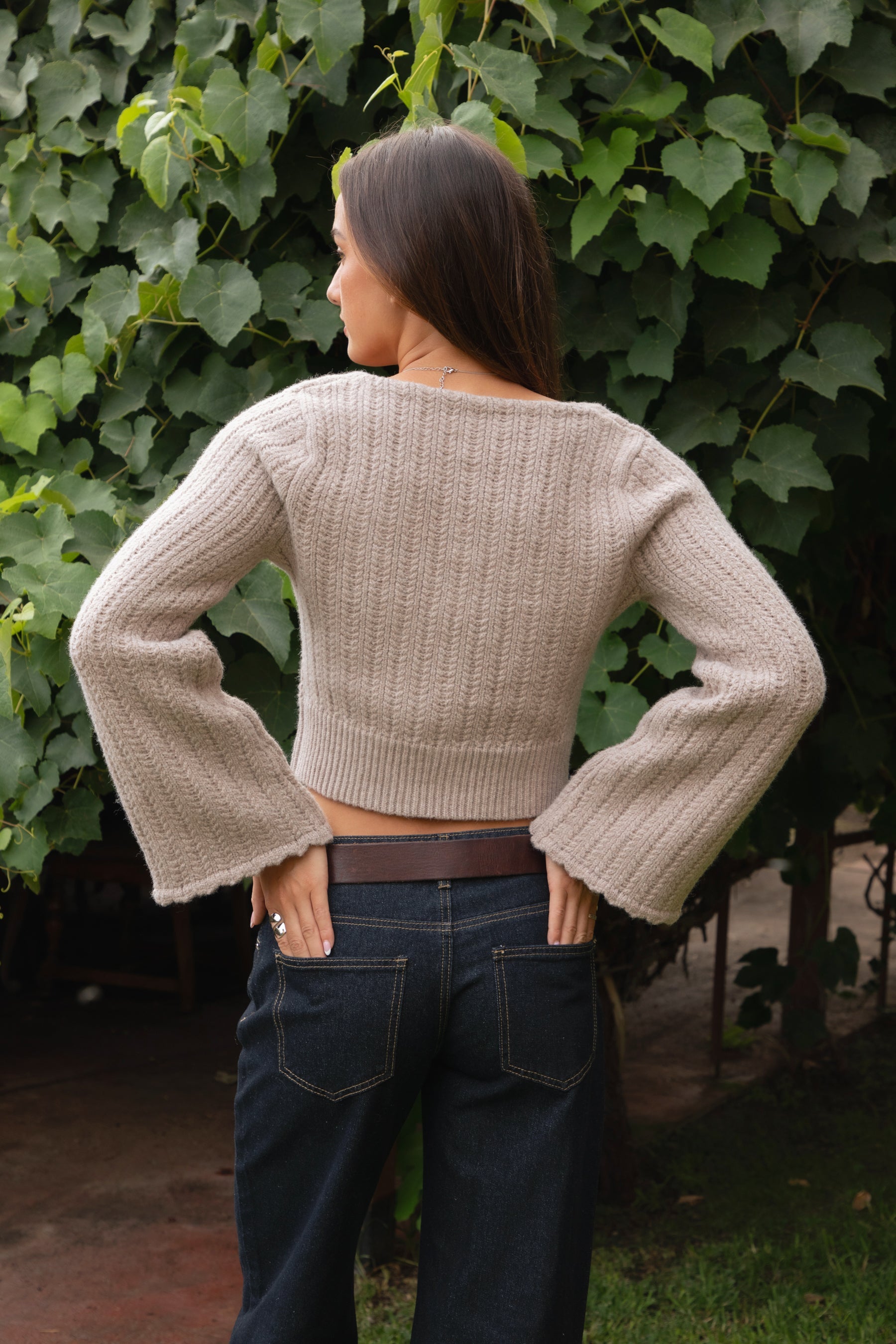 Woman wearing TIC TOC TV31638 HEARTH marled taupe cable knit button down cardigan with scalloped hem and relaxed bell sleeves with dark denim pants  against a green leafy background.