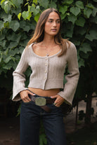 Woman wearing TIC TOC TV31638 HEARTH marled taupe cable knit button down cardigan with scalloped hem and relaxed bell sleeves with dark denim pants  against a green leafy background.