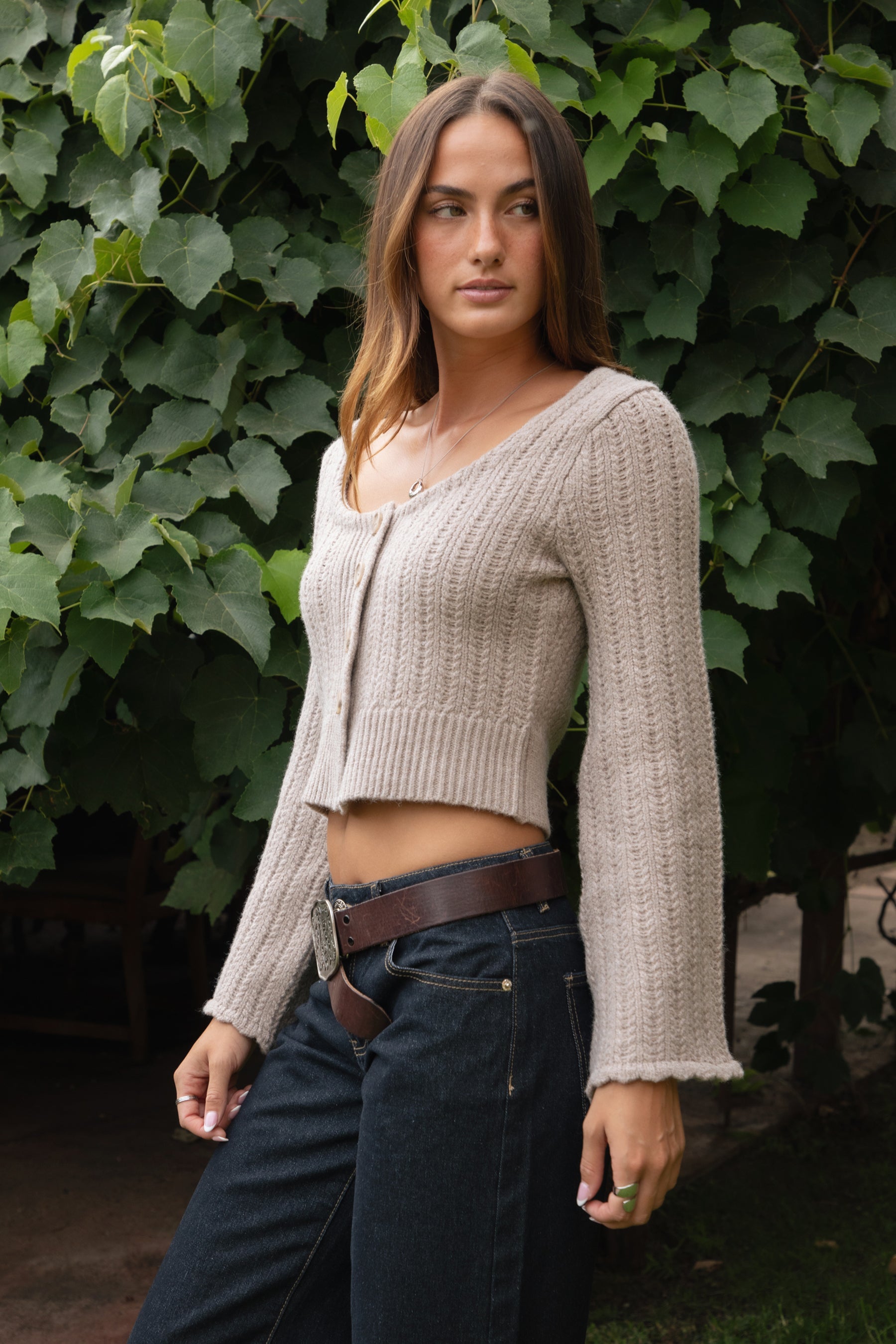 Woman wearing TIC TOC TV31638 HEARTH marled taupe cable knit button down cardigan with scalloped hem and relaxed bell sleeves with dark denim pants  against a green leafy background.