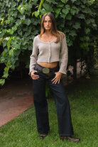 Woman wearing TIC TOC TV31638 HEARTH marled taupe cable knit button down cardigan with scalloped hem and relaxed bell sleeves with dark denim pants  against a green leafy background.