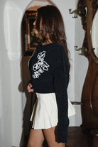 Woman wearing TIC TOC TW31732 WILLOWEN black embroidered knit button down cardigan with white western embroidery paired with a white mini skort in an indoor setting. 